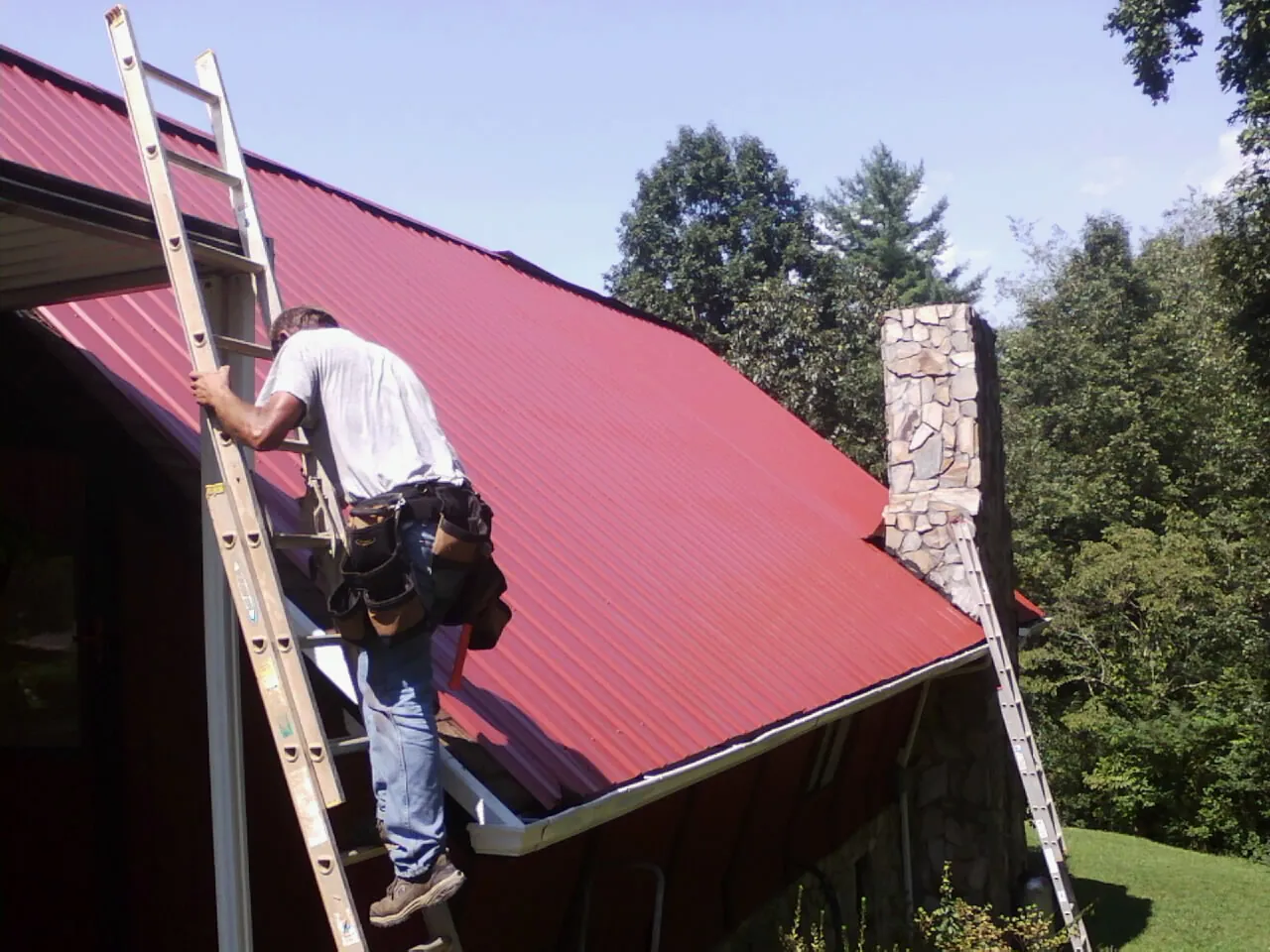 Roofer on a ladder beside a red metal roof and stone chimney — Soffit & Fascia Repair by Roofing Brothers in Bonfield